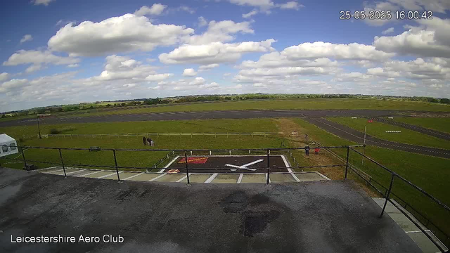 A wide view of an airfield landscape under a partly cloudy sky. In the foreground, there is a helicopter pad with a white helicopter parked to the left. A black and white helicopter landing pad with a white "H" is centered. A couple of people are near the grass area to the left of the helipad. The grass extends outward, with a fence separating the airfield from the distant landscape. The horizon shows open fields and a distant treeline. The image includes a timestamp in the top right corner showing "25-05-2025 16:00:42" and the bottom left corner has the text "Leicestershire Aero Club."