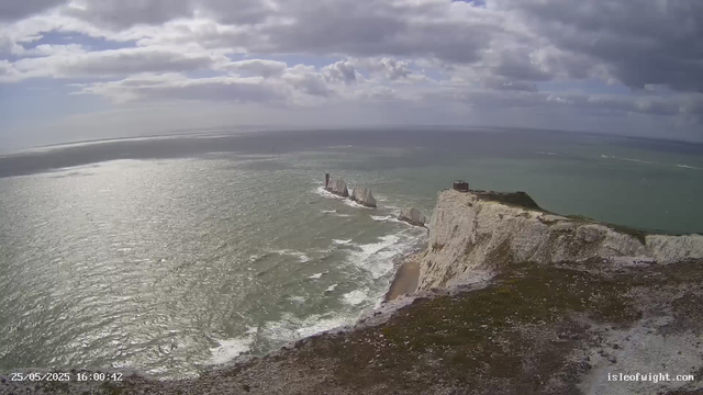 Aerial view of white chalk cliffs with grassy tops along the shoreline, with the sea stretching out to the horizon. Dark clouds cover the sky, casting shadows over the water, which reflects the overcast light. The cliffs feature a small building at the edge, and a series of rocky sea stacks extend into the ocean. Waves gently wash against the cliffs and rocks below. The timestamp in the bottom left corner reads "25/05/2025 16:00:42," and the bottom right corner shows "isleofwight.com."