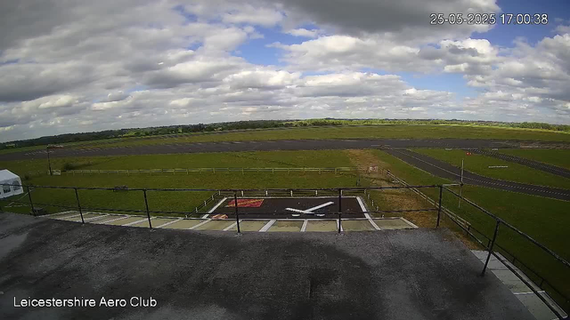 A view of an airfield with a helipad in the foreground, marked with a large white helicopter symbol and a red square, surrounded by a black border. There is a white tent on the left side. The grassy airfield extends into the distance with a runway visible on the right side. The sky is partly cloudy with patches of blue. The image is timestamped "25-05-2025 17:00:38" in the top right corner, and "Leicestershire Aero Club" is written in the bottom left corner.