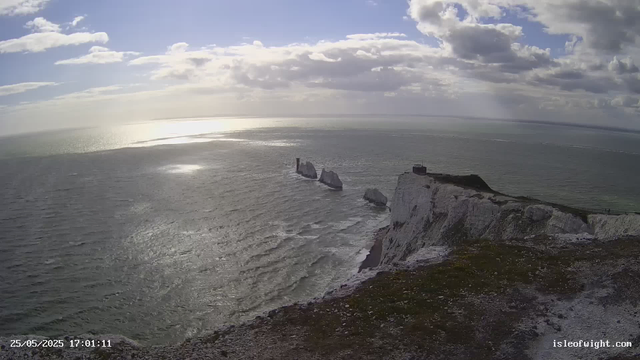 A view of white chalk cliffs overlooking the ocean, with a small building or lighthouse on top of the tallest cliff. Several large sea stacks rise from the water near the base of the cliffs, extending into the sea. The sky is partly cloudy with sunlight reflecting off the ocean surface. The timestamp indicates the photo was taken on May 25, 2025, at 17:01.