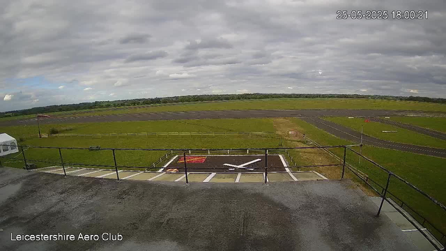 A view from a webcam showing an airfield with a helipad in the foreground and a runway extending into the distance under a cloudy sky. The image is marked with the date and time as 25-05-2025 at 18:00:21, and the location is identified as Leicestershire Aero Club. There are some grassy areas and a few small structures around the helipad, with a fence along the edge of the airfield.