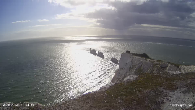 A view from a webcam showing white chalk cliffs along the coastline, with the ocean stretching out towards the horizon. The sky is partly cloudy, with the sun shining brightly, reflecting off the water. The cliffs are rugged and steep, with some patches of grass at the top. The photo was taken in the evening, as indicated by the timestamp "25/05/2025 18:01:10."