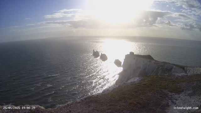 A view of the ocean at sunset or sunrise with the sun low on the horizon, casting bright reflections on the water. There are white chalk cliffs on the right side of the image with a small building or lighthouse on top. In the distance, dark rocks or sea stacks rise from the water near the shoreline. The sky is partly cloudy with a mix of light and dark clouds. The image also includes a timestamp in the lower left corner and a website URL in the lower right corner.