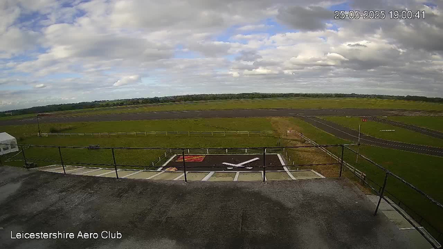 An outdoor scene of an airfield under partly cloudy skies. The foreground shows the top of a building or observation deck with safety railings. In the middle of the image, there is a helicopter landing pad with a white helicopter symbol painted on it. The landscape extends into grassy fields and taxiways, with a runway visible in the distance. A small tent can be seen on the left side. The image is timestamped "25-05-2025 19:00:41" at the top right corner, and "Leicestershire Aero Club" is written at the bottom left corner.