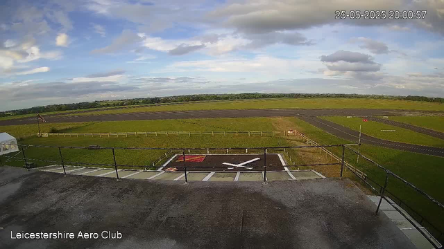 A view from a webcam at Leicestershire Aero Club showing a vast open grassy airfield with a runway stretching across the landscape. The sky is partly cloudy with patches of blue, and the timestamp indicates the date and time as 25th May 2025 at 8:00 PM. In the foreground, there is a small section of a rooftop with safety railings, and a helipad with a white 'H' marked on it. The scene appears calm and quiet with no aircraft or people visible.