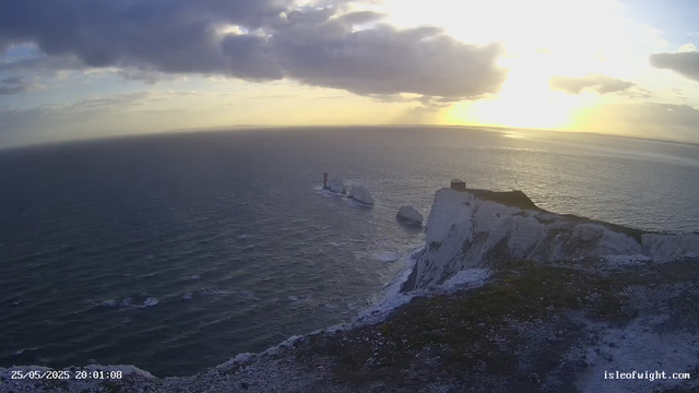 A panoramic view of white chalk cliffs extending into the ocean at sunset. The sky is partly cloudy with the sun low on the horizon, casting a warm glow over the water and landscape. There is a lighthouse and a small building on the highest point of the cliffs. Waves gently lap against the base of the cliffs. The scene has a serene, natural beauty with a mix of dark clouds and golden sunlight. The timestamp reads "25/05/2025 20:01:08" and the bottom right corner displays "isleofwight.com."