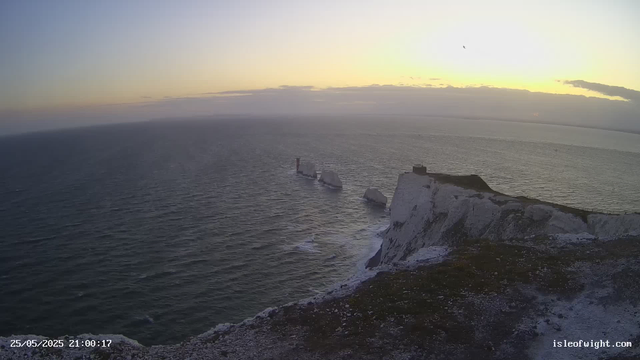 A view from a webcam showing white chalk cliffs along a coastline at sunset. The cliffs extend into the sea with some rocks visible at the base. The sky is partly cloudy with the sun setting, casting a warm glow on the horizon. A single bird is flying in the sky. The timestamp shows the date and time as 25/05/2025 at 21:00:17, and the website is isleofwight.com.