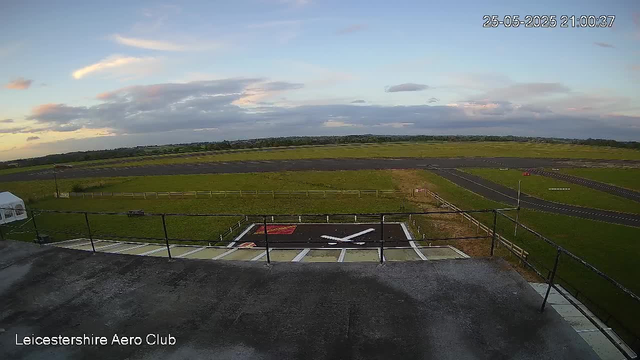 A view from a webcam at Leicesterhire Aero Club showing an open sky with scattered clouds during sunset. In the foreground, there is a helicopter landing pad with a white helicopter parked on it. The landing pad is surrounded by a safety fence, and a small white tent can be seen on the left side. In the background, there are grassy fields and a runway extending into the distance, with a few distant trees on the horizon. The timestamp in the top right corner reads "25-05-2025 21:00:37."