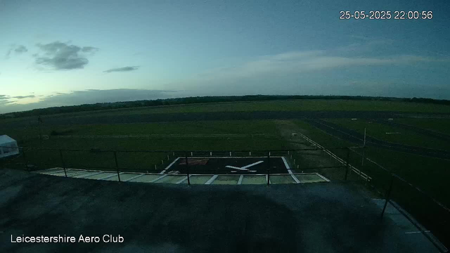 An early evening view from a webcam at Leicestershire Aero Club shows a helipad with a large white helicopter symbol. The surrounding landscape features green fields, some trees in the distance, and a partly cloudy sky with the current date and time displayed as 25-05-2025 22:00:56.