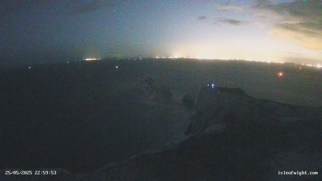 A dark, nighttime scene with a distant horizon illuminated by bright lights, possibly from a city or fireworks. The foreground shows faint outlines of land or rocks, and a small, blurred object is present in the lower part of the image. The timestamp in the bottom left indicates the date and time as May 25, 2025, at 22:59:53, and there is a website URL "isleofwight.com" in the bottom right corner.