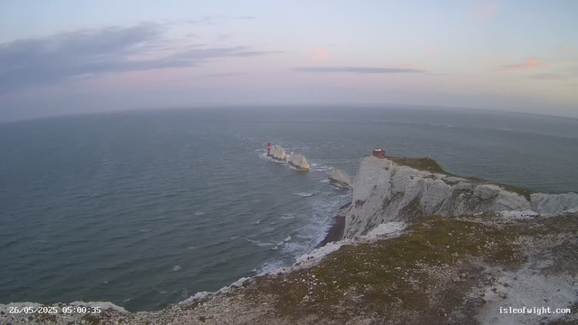 A view from a webcam showing white chalk cliffs extending into the sea, with several large rocks and a red and white lighthouse standing on one of the rocks offshore. The sky has soft pastel colors, indicating either dawn or dusk, and the ocean appears calm with gentle waves. The image is timestamped "26/05/2025 05:00:35" and has the website "isleofwight.com" in the bottom corner.