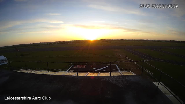 A sunrise over a flat, open landscape with a sky filled with light clouds. The horizon shows distant trees and fields. In the foreground, there is a helipad with markings and a metal railing around its edge, with a small white tent on the left side. The image is timestamped "26-05-2025 05:00:59" and labeled "Leicestershire Aero Club."