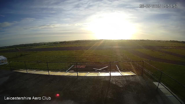 A wide view of an open grassy airfield at sunrise or sunset, with a low sun near the horizon, casting a bright glow across the landscape. The sky has scattered clouds, and the date and time are displayed as 26-05-2025 05:59:57. The foreground shows a paved area with a railing, and the words "Leicestershire Aero Club" are written in the lower left corner.