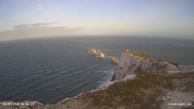 An aerial view of white chalk cliffs extending into the sea, with a lighthouse and a small building on top. Several large rocks are visible in the water near the cliffs, and the horizon shows a calm sea with a few distant boats or ships. The sky is partly cloudy with soft light, indicating early morning or late afternoon. The timestamp in the corner shows the date as May 26, 2025, and the time as 06:00:14.