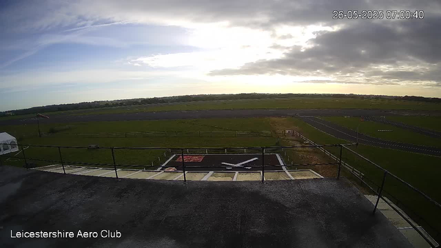 A wide view of an airfield during early morning with the sky partly cloudy. In the foreground, there is a helipad with a large white bird symbol painted in the center. The airfield is surrounded by grassy fields and a runway stretching into the distance. There is a fence and a small structure on the left side. The timestamp at the top right indicates the date and time as May 26, 2025, at 7:00:40 AM. The text "Leicestershire Aero Club" is displayed in the bottom left corner.