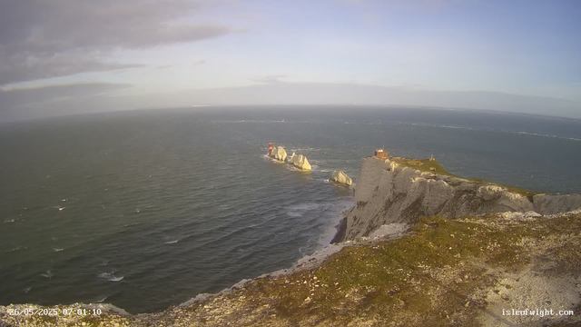 A panoramic view of a rugged coastal landscape with tall white chalk cliffs leading down to the sea. Several large rocks are visible offshore, with a red lighthouse perched on one of the rocks. There is a small building with an antenna on the top of the cliff. The water is slightly choppy with small waves, and the sky above has scattered clouds with patches of blue. The scene appears to be taken early in the morning or late afternoon based on the lighting.
