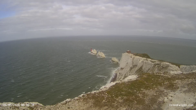 A view from a webcam shows white chalk cliffs descending into the ocean below. Several large rocks are visible in the water, with a red and white lighthouse perched on one of the rocks and a small building on top of the cliff. The water is slightly choppy, and the sky is mostly cloudy with patches of blue. The timestamp in the corner indicates the image was captured on May 26, 2025, at 8:00 AM.