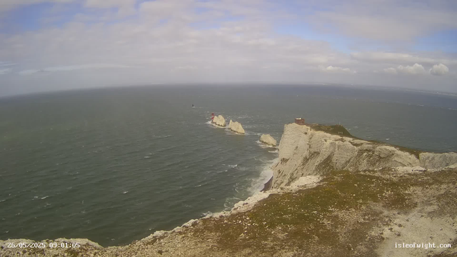 An aerial view of white chalk cliffs along the coast with a small building on top. The cliffs stretch into the ocean, where several rock formations and a lighthouse are visible in the distance. The sky is partly cloudy with patches of blue. The image includes a timestamp of "26/05/2025 09:01:05" and the website "isleofwight.com" in the bottom corners.
