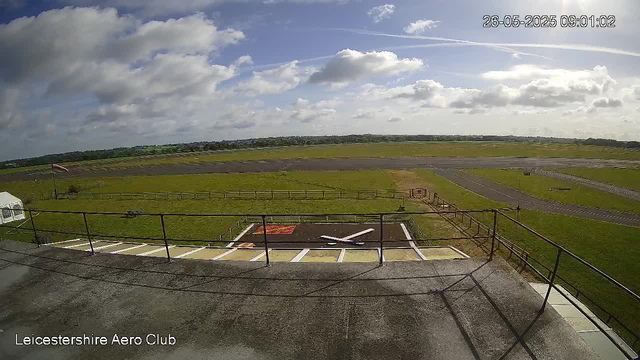 A view from a webcam at LeicesterShire Aero Club showing a partly cloudy sky with scattered clouds and sunlight illuminating the scene. The image captures a grassy tarmac area with a small helicopter landing pad, an aircraft or drone on the ground, and surrounding open fields with a runway in the distance. There is a railing in the foreground and the timestamp in the top right corner indicates the date and time as 26th May 2025 at 09:01:02.