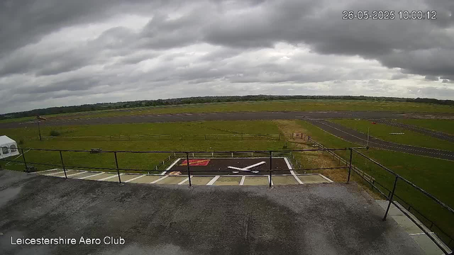 An image from a webcam shows an overcast sky with dark clouds above an empty airfield. In the foreground, there is a helicopter landing pad marked with a large white "H" and a red square nearby. The airfield has a grassy area surrounding the paved runway and taxiways. A small tent is visible on the left side. The timestamp in the top right corner reads "26-05-2025 10:00:12," and the bottom left corner indicates "Leicestershire Aero Club." The overall scene appears calm with no aircraft or people visible.