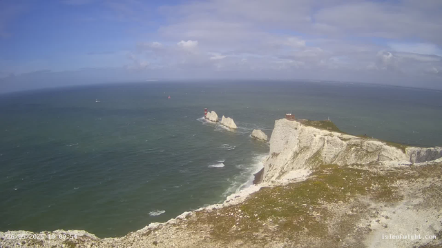 A panoramic view of white chalk cliffs along a rugged coastline, with a lighthouse and a small building perched on one of the cliffs. The ocean stretches out into the distance with sailboats and a lighthouse in sight. The sky is partly cloudy with patches of blue. The scene captures a broad sweep of the coastline with waves crashing against the rocks below.