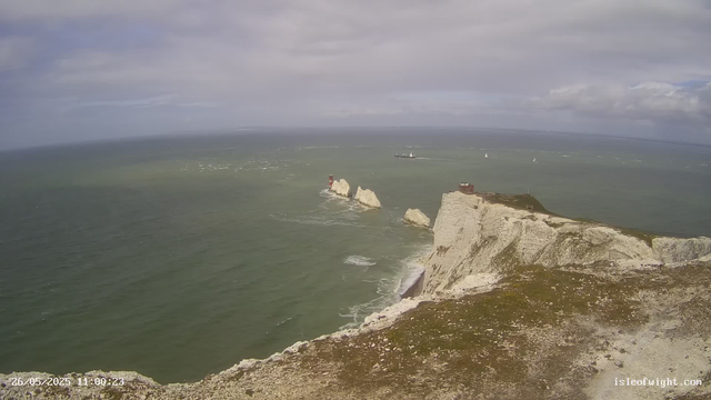 A view of white chalk cliffs along a coastline extending into the distance, with a lighthouse on the far left and another in the middle, both situated on separate rock formations in the sea. Several sailboats and ships are visible on the water under a partly cloudy sky. The timestamp indicates the date and time as May 26, 2025, at 11:00:23.