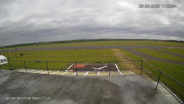 A view from a webcam at Leicesterhire Aero Club showing an overcast sky with dark grey clouds. The scene includes a small parking area with marked parking spaces, a helipad with a white "H" in the foreground, and a windsock on a pole to the left. The runway and taxiways stretch into the distance, surrounded by green grass. The timestamp in the top right corner reads "26-05-2025 11:00:34." The bottom left corner displays the text "Leicestershire Aero Club."