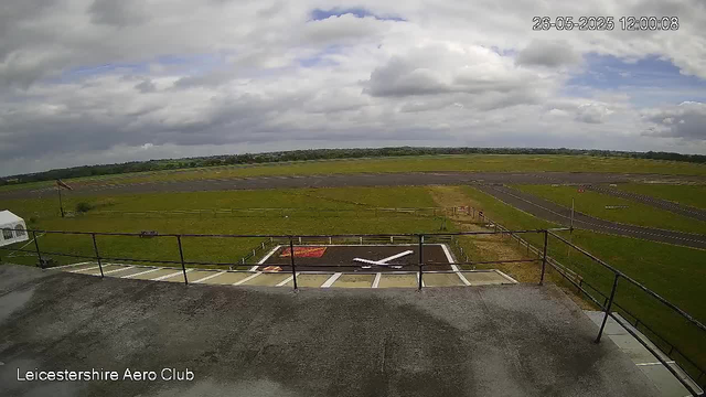 A view from a webcam at Leicesterhire Aero Club shows an open sky with scattered clouds. In the foreground, there is a concrete platform with a railing, and a helicopter parking area with a black and white helicopter silhouette painted on the ground. The surrounding area is a grassy field with a runway visible in the distance. The timestamp in the upper right corner reads "26-05-2025 12:00:08."