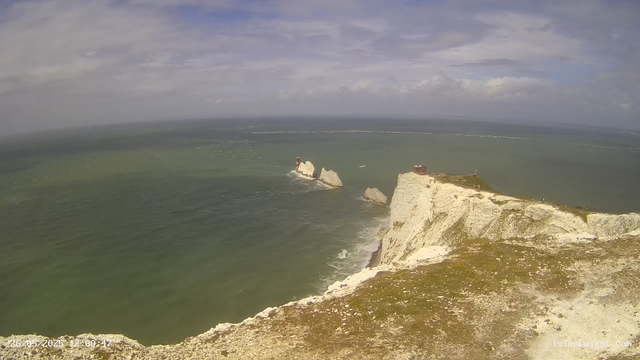 A coastal view from a high vantage point showing white chalk cliffs extending into the ocean with a lighthouse on one of the rocks. The sea is greenish with gentle waves, and the sky is partly cloudy with patches of blue.
