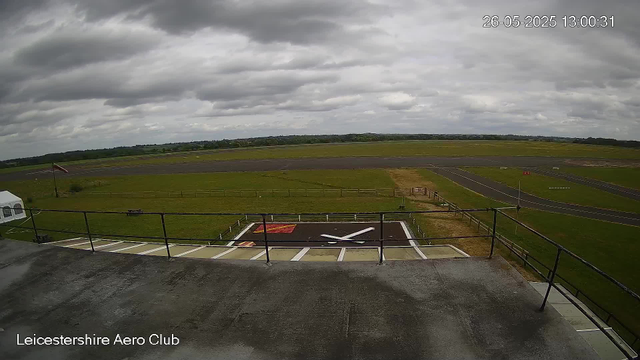 The image shows an overcast view from a webcam at Leicestershire Aero Club. In the foreground, there is a large clock painted on the ground, with its white hour and minute hands pointing to approximately 2:09. A railing surrounds the clock area. Beyond the clock, there is a grassy area with a small windsock on a pole to the left. Extending further, there are runways and taxiways for aircraft, with some small markings and lights visible. The sky above is filled with dark, dense clouds, creating a cloudy and slightly gloomy atmosphere. The timestamp in the upper right corner reads "26-05-2025 13:00:31."