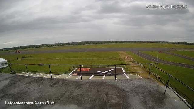 A panoramic view from a webcam at Leicestershire Aero Club, showing an overcast sky with thick gray clouds. In the foreground, there's a helipad with a white helicopter outline and a red square, surrounded by a white and black border. A small portion of the rooftop and railing are visible at the bottom of the image. Beyond the helipad, there are grassy fields, taxiways, and runways stretching into the distance. A weathered wooden fence runs along the edge of the grassy area, and a red flag is flying on a pole near the left side. The overall scene is calm and quiet.