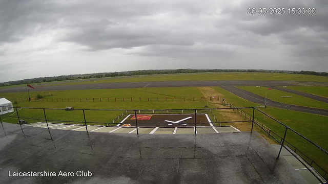 A view from a webcam at Leicestershire Aero Club showing a cloudy sky over an airfield. In the foreground, there is a helicopter landing pad with a large white "X" marked on it, and a small red and white rectangular marking nearby. The airfield has grassy areas, runways, and taxiways stretching into the distance. There is a small tent on the left side near the fence, and a flagpole with a red flag. The timestamp indicates the date as May 26, 2025, at 3:00 PM. The overall scene is calm and overcast.