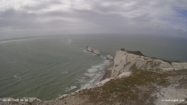 A view of white chalk cliffs overlooking the ocean on a cloudy day. There are three prominent sea stacks rising from the water near the cliffs. A lighthouse is situated on the edge of the highest cliff. The coastline extends into the distance under a partly cloudy sky. The timestamp reads "26/05/2025 15:00:21" and the website "islefowight.com" is visible in the bottom right corner.