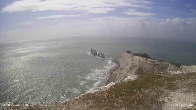 A view of the ocean with white cliffs along the shoreline. There are large rock formations and a lighthouse in the distance on the cliffs. The sky is mostly cloudy with patches of blue, and the water appears slightly choppy. The image is timestamped "26/05/2025 16:00:25" at the bottom left corner.