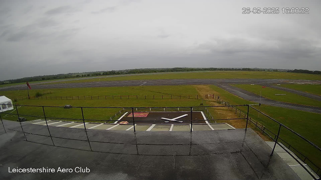 A view from a webcam at Leicesterhire Aero Club shows an overcast sky with gray clouds. In the foreground, there is a helipad with a large white "X" on a black background. The surrounding area includes a grassy field with a small fenced area, a flagpole with a red flag, and a white tent on the left side. Extending into the background, the scene reveals a paved area with taxiways and runways. The overall atmosphere appears calm and slightly damp, possibly after recent rain. The timestamp in the top right corner reads "26-05-2025 16:00:32".