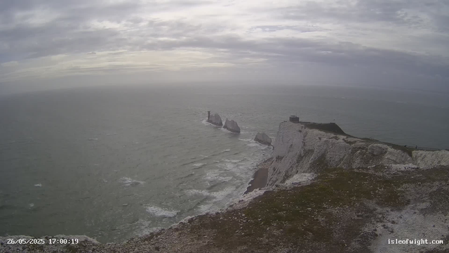 A view of white chalk cliffs along the coastline with a lighthouse on the edge. The ocean waves crash against the base of the cliffs, and several sea stacks protrude from the water. The sky is overcast with gray clouds, creating a moody atmosphere.