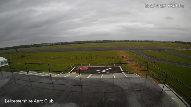 An overcast sky looms above a runway and grassy airfield at Leicester Aero Club. In the foreground, there's a rooftop area with metal railings and a helipad marked with a large white "X." The runway extends into the distance, bordered by green fields and some distant trees. The timestamp in the top right corner shows the date as May 26, 2025, at 17:01.