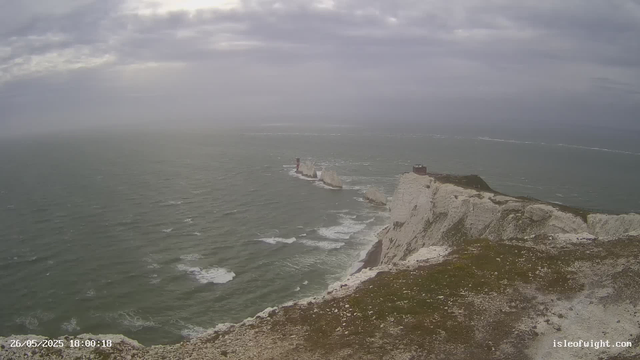 A view of white chalk cliffs along the coastline, with the sea crashing against their bases. A lighthouse stands on a small outcrop on the cliffs, and a building is visible further along the cliff edge. The sky is overcast with grey clouds, and the ocean appears choppy with waves. The scene has a moody, cloudy atmosphere.