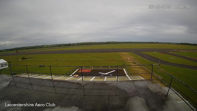 An empty helicopter landing pad with a white and red marking, is situated on a rooftop. A black railing surrounds the area, and a windsock pole is visible on the left. Beyond the rooftop, there is a wide grassy field leading to a runway, under a cloudy, overcast sky. The image includes a timestamp in the top right corner and the label "Leicestershire Aero Club" at the bottom left.