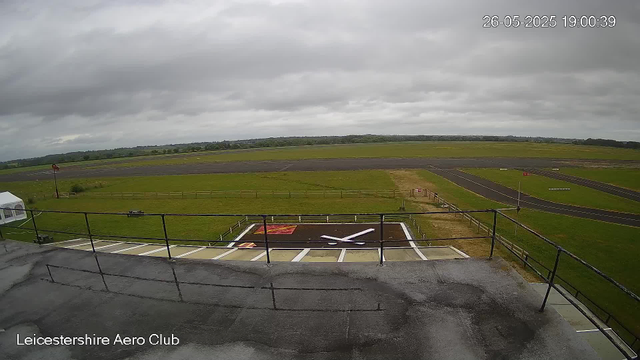 A view from a webcam showing an overcast sky above an airfield. In the foreground, there is a parking area with designated spaces and a helipad marked with a large white "H" on a dark surface. There is a small tent and a windsock near the helipad. Surrounding the airfield are grassy fields and a taxiway extending into the distance, with a few runway markings visible. The image is labeled "Leicestershire Aero Club" in the bottom left corner, and the timestamp in the top right corner reads "26-05-2025 19:00:39."