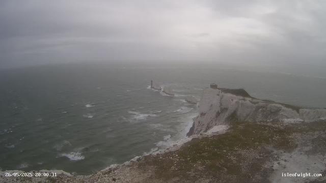 A view of white chalk cliffs extending into the ocean under a cloudy sky. The cliffs have patches of green vegetation, and there is a small building or structure at the edge of the highest point of the cliffs. The sea below is slightly choppy with white surf near the base of the cliffs. There are rock formations in the water further out from the shore. The timestamp on the image reads "26/05/2025 20:00:31" and the website "isleofwight.com" is visible in the bottom right corner.