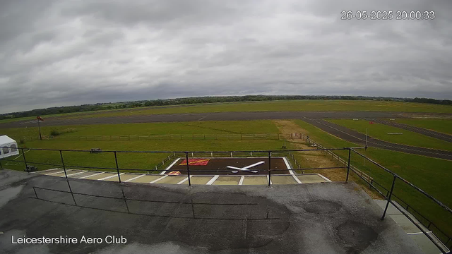 A view from a webcam at Leicestershire Aero Club shows a cloudy sky over a grassy airfield with runways extending into the distance. In the foreground, there is a helipad with a large white "X" mark, surrounded by a safety railing. To the left, a small white tent and a windsock are visible. The landscape is flat with open fields and a few trees, and the time and date displayed in the upper right corner are May 26, 2025, at 8:00 PM and 33 seconds.