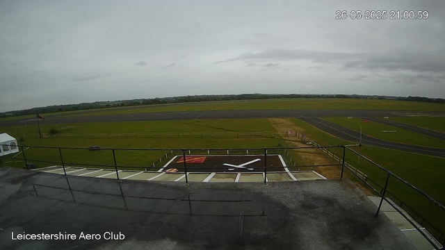 An overcast sky overlooking the runway and grassy fields of Leicestershire Aero Club. The image shows a large clock on the ground, some fencing, a windsock, and a small tent on the side. The scene appears to be captured from an elevated vantage point on a building or observation deck. The date and time stamp in the top right corner reads "26-05-2025 21:00:59."