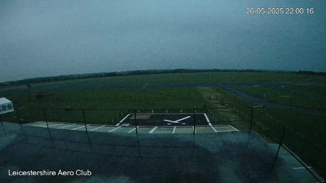 An empty helicopter landing pad with a large white 'X' marking in the center, surrounded by a black border and white dashed lines. The landing pad is situated on a rooftop with a metal railing, overlooking a grassy field and distant horizon under a darkening evening sky. The timestamp in the top right corner shows "26-05-2025 22:00:16", and the bottom left corner indicates the location as "Leicestershire Aero Club".
