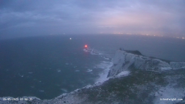 A view from a webcam at dusk shows a rugged coastline with tall, white chalk cliffs meeting the sea. The ocean has waves crashing against the rocks. There are red and white navigation lights on the cliff edge, indicating a lighthouse or beacon. In the background, there are faint city lights along the horizon under a cloudy sky. The timestamp shows the date and time as 26/05/2025 at 22:00:35, and the URL is isleofwight.com.
