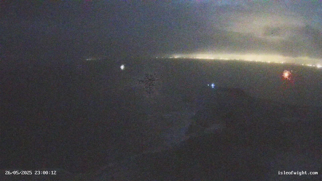 A dark nighttime scene viewed through a webcam, featuring a faint horizon line with light coming from distant city lights or stars. The sky is cloudy, and there are some small, distant bright spots that could be stars or navigation lights. The image includes a timestamp at the bottom left corner, indicating the date and time as May 26, 2025, at 23:00:12, and a website URL "isleofwight.com" at the bottom right corner.
