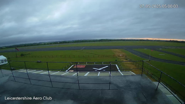 An outdoor view of an airport runway and grassy area, captured from a webcam at the Leicestershire Aero Club. The sky is overcast with dark clouds, and a flag is visible on the left side. In the foreground, there is a helipad marked with a white 'H' and surrounded by a safety fence. The timestamp indicates it is early morning. The photo conveys a quiet, cloudy day at the airfield.
