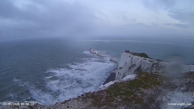 A view of white chalk cliffs extending into the ocean with waves crashing against the base. There are two prominent lighthouse structures on the cliffs, one at the far end and the other near the middle. The sky is cloudy, giving the scene a moody, overcast atmosphere. A timestamp in the bottom left corner indicates the date and time as May 27, 2025, at 5:00:33 AM, and there is a website URL "isleofwight.com" in the bottom right corner.