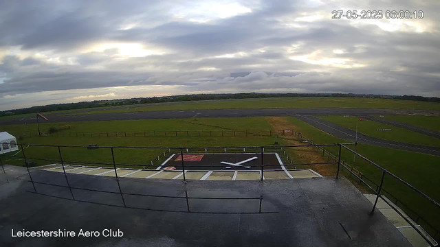 An outdoor view from a webcam at Leicesterhire Aero Club, showing a runway and grassy area under a cloudy sky with patches of sunlight. The image is timestamped 27-05-2025 06:00:10. In the foreground, there is a balcony with metal railings and a small helicopter landing pad marked with a white "X" and a red square.