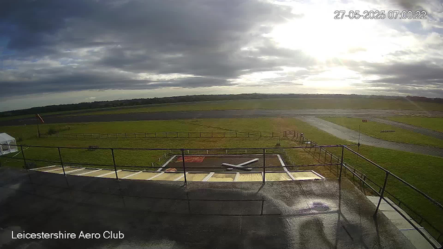 A view from a webcam showing an outdoor scene at Leicester Aero Club. In the foreground, there is a helicopter landing pad with some equipment and a small tent on the left side. The pad is fenced, and beyond it, there are green grassy fields and a paved runway area. The sky is partly cloudy with the sun shining through the clouds, creating a bright spot in the sky. The timestamp on the image indicates it was taken on May 27, 2025, at 7:00 AM.